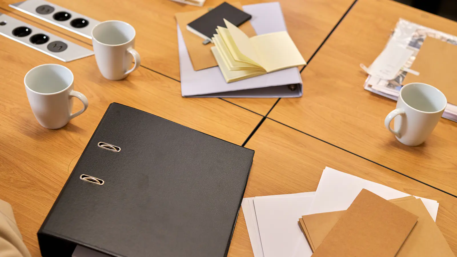 Meeting table with a ring binder, loose forms, a notebook and two cups